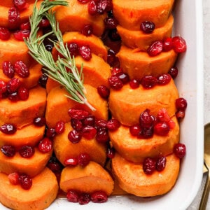 A white casserole dish filled with neatly arranged sliced sweet potatoes, topped with cranberries and garnished with a sprig of rosemary.