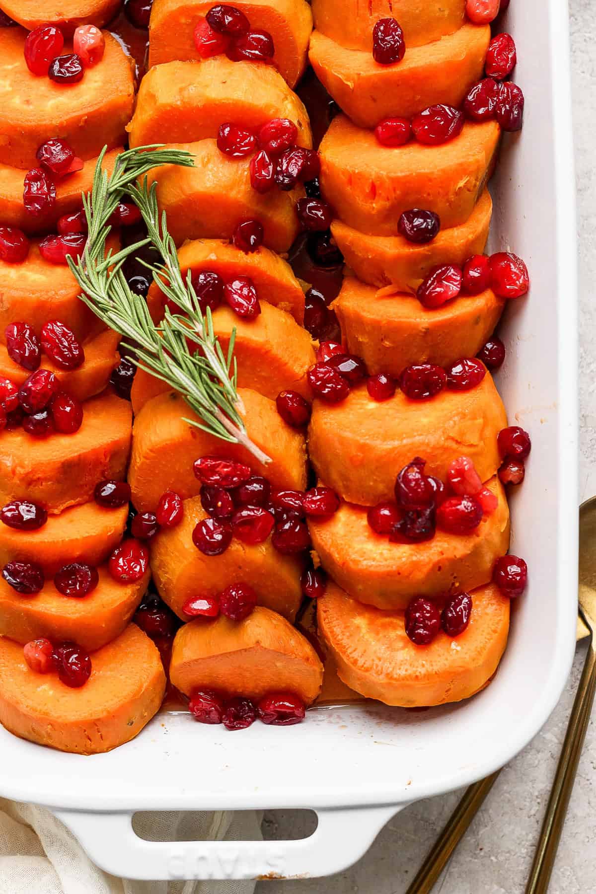 A white casserole dish filled with neatly arranged sliced sweet potatoes, topped with cranberries and garnished with a sprig of rosemary.