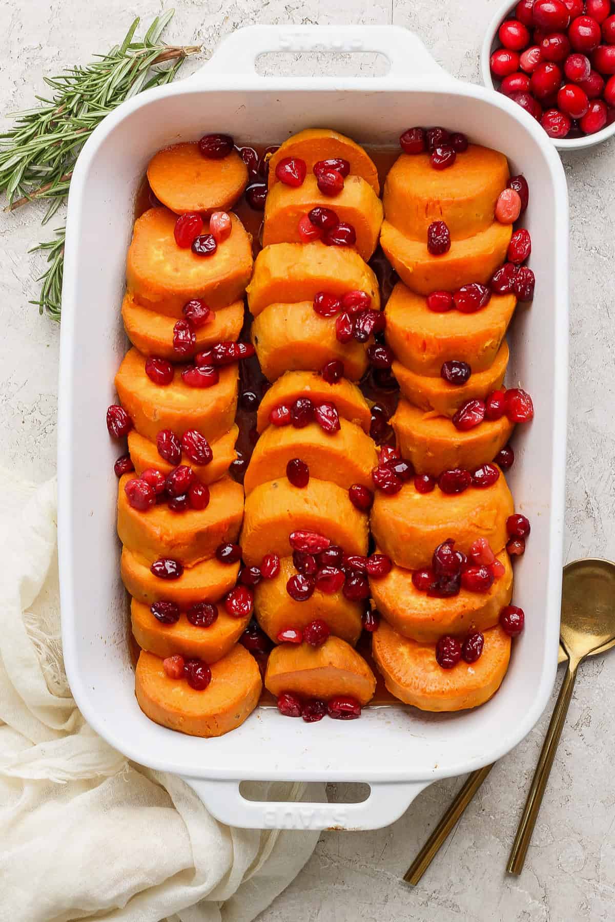 A white baking dish filled with sliced sweet potatoes topped with cranberries. A sprig of rosemary, a bowl of cranberries, and two gold utensils are placed beside the dish.