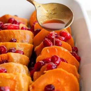 A spoon drizzles liquid over sliced sweet potatoes topped with cranberries in a rectangular white baking dish.