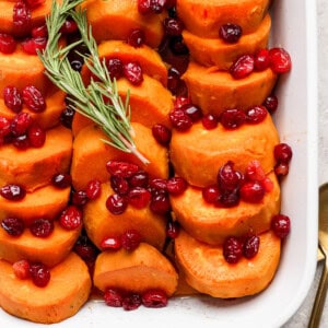 Sliced sweet potatoes topped with cranberries and garnished with a sprig of rosemary, served in a white dish.