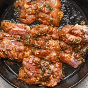 Raw marinated chicken thighs in a dark cast iron skillet, ready to be cooked. The marinade includes herbs and a reddish sauce. The skillet rests on a light, textured surface.