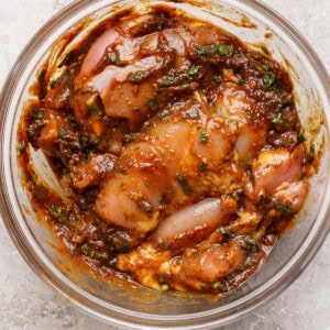 A clear glass bowl containing marinated pieces of raw chicken mixed with herbs and spices in a thick, reddish-brown sauce.