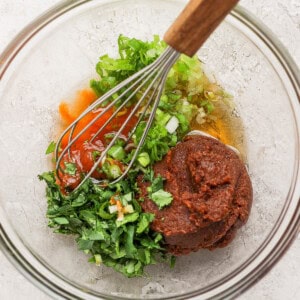 A glass bowl containing chopped green herbs, red sauce, a thick brown paste, and a whisk with a wooden handle on a light-colored surface.