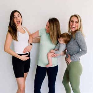 Three women standing, two happily touching their stomachs. One is holding a baby. They are smiling in casual athletic wear against a plain background.