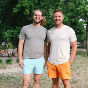 Two people smiling and standing outdoors in a park, wearing casual t-shirts and shorts, surrounded by greenery.