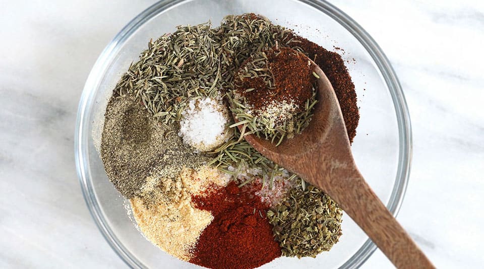 A clear bowl filled with assorted spices and herbs, including ground pepper, paprika, salt, oregano, and thyme, stirred with a wooden spoon.