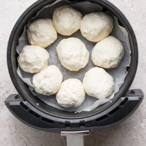 An air fryer basket lined with parchment paper holds eight uncooked dough balls in a circular arrangement.