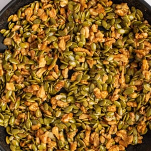 A pan filled with a mix of nuts and seeds, predominantly featuring green pumpkin seeds and walnuts, with a light coating, likely for roasting.