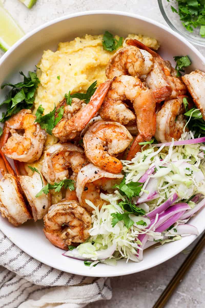 A bowl of grilled shrimp with creamy grits, garnished with chopped cilantro and served with a side of shredded cabbage and red onion.