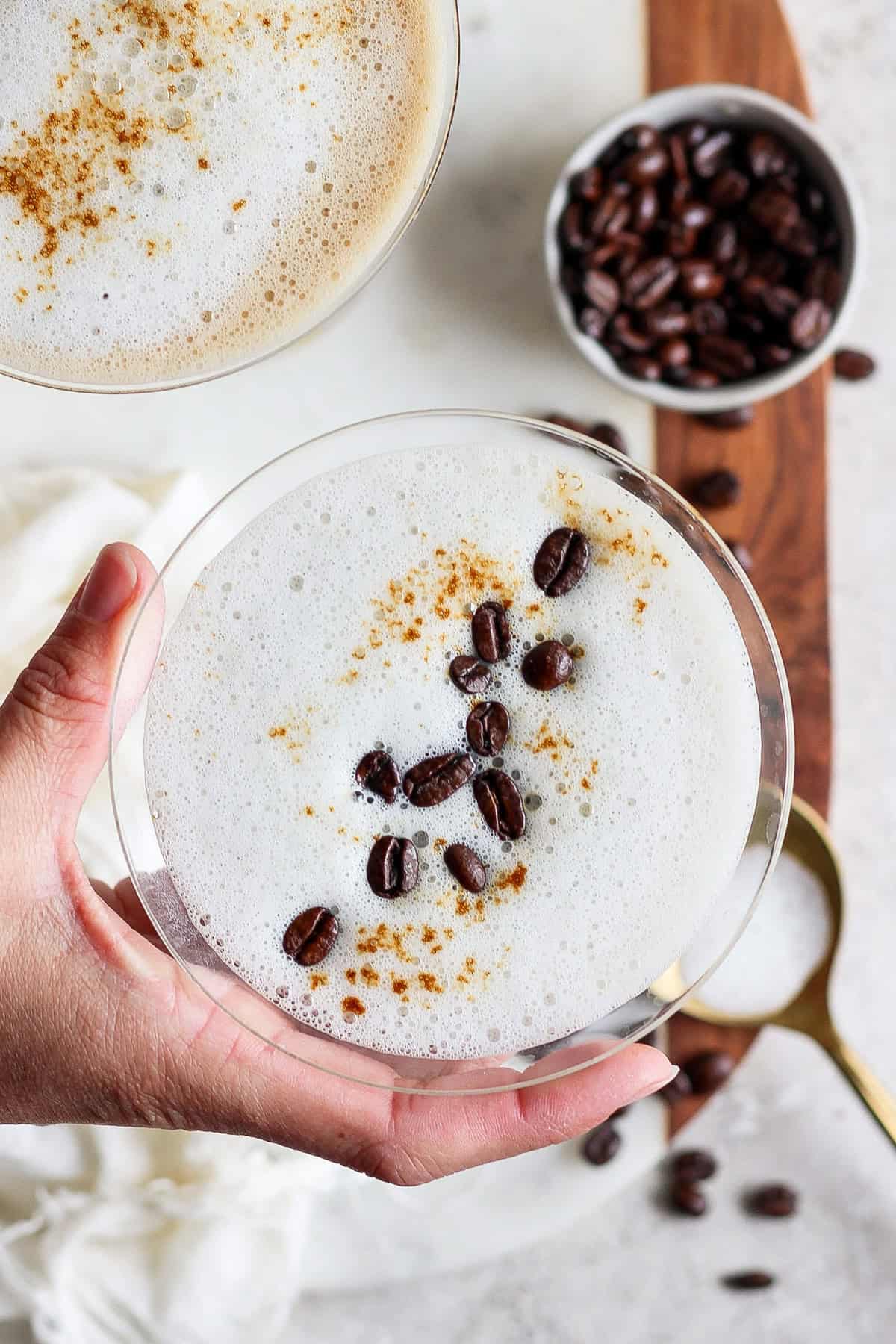 A hand holding a frothy coffee drink in a glass, garnished with coffee beans and cinnamon. Another glass and a bowl of coffee beans are visible in the background.