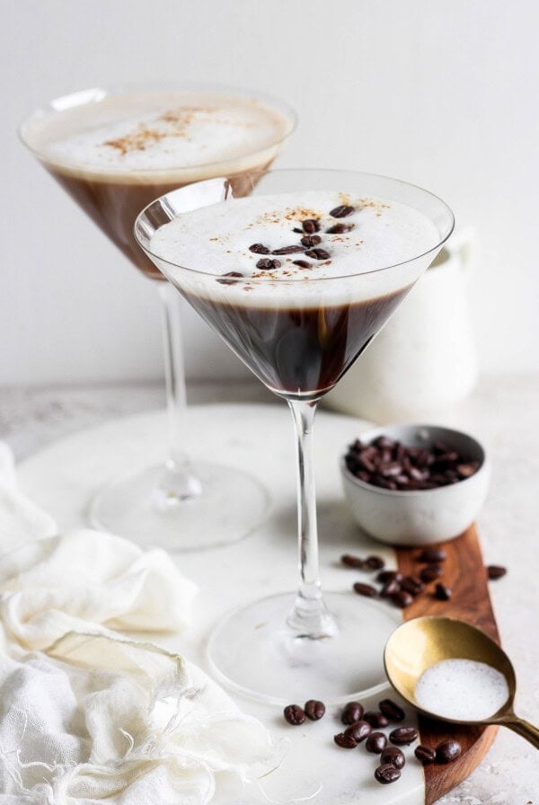 Two espresso martinis in martini glasses, garnished with coffee beans, on a wooden board with a small bowl of coffee beans and a brass spoon nearby. A white cloth is draped on the left.