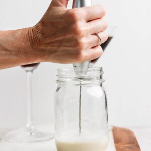 A hand holds a milk frother in a glass jar on a marble board, creating foam. Two stem glasses are in the background.