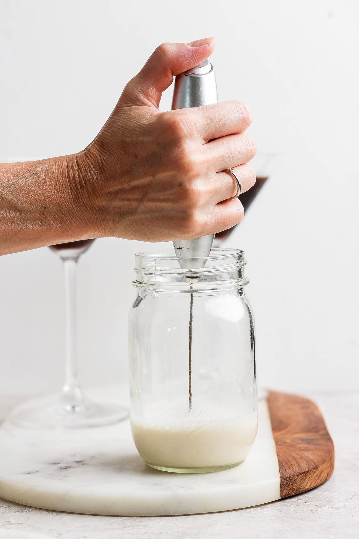 A hand holds a milk frother in a glass jar on a marble board, creating foam. Two stem glasses are in the background.