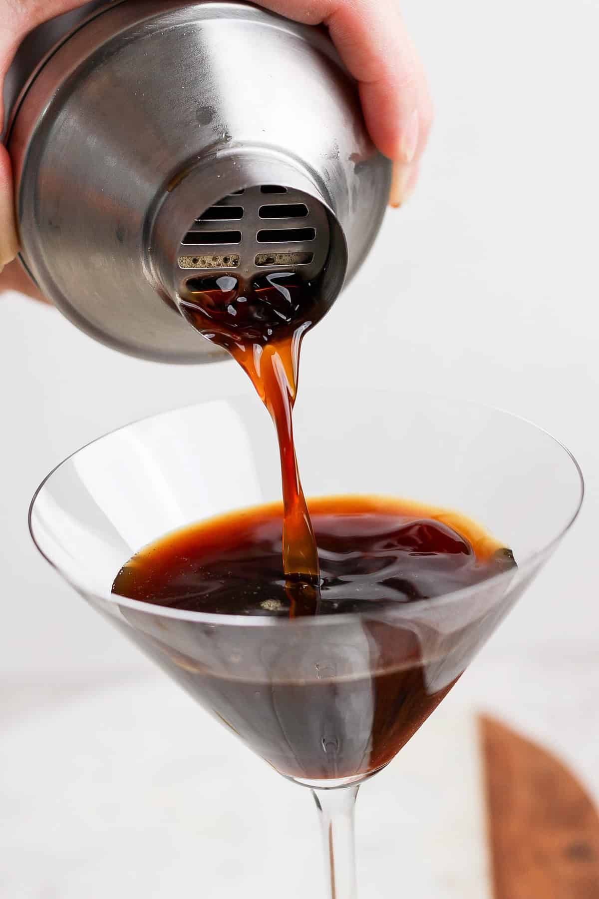 A hand pouring a dark liquid from a metal shaker into a martini glass.