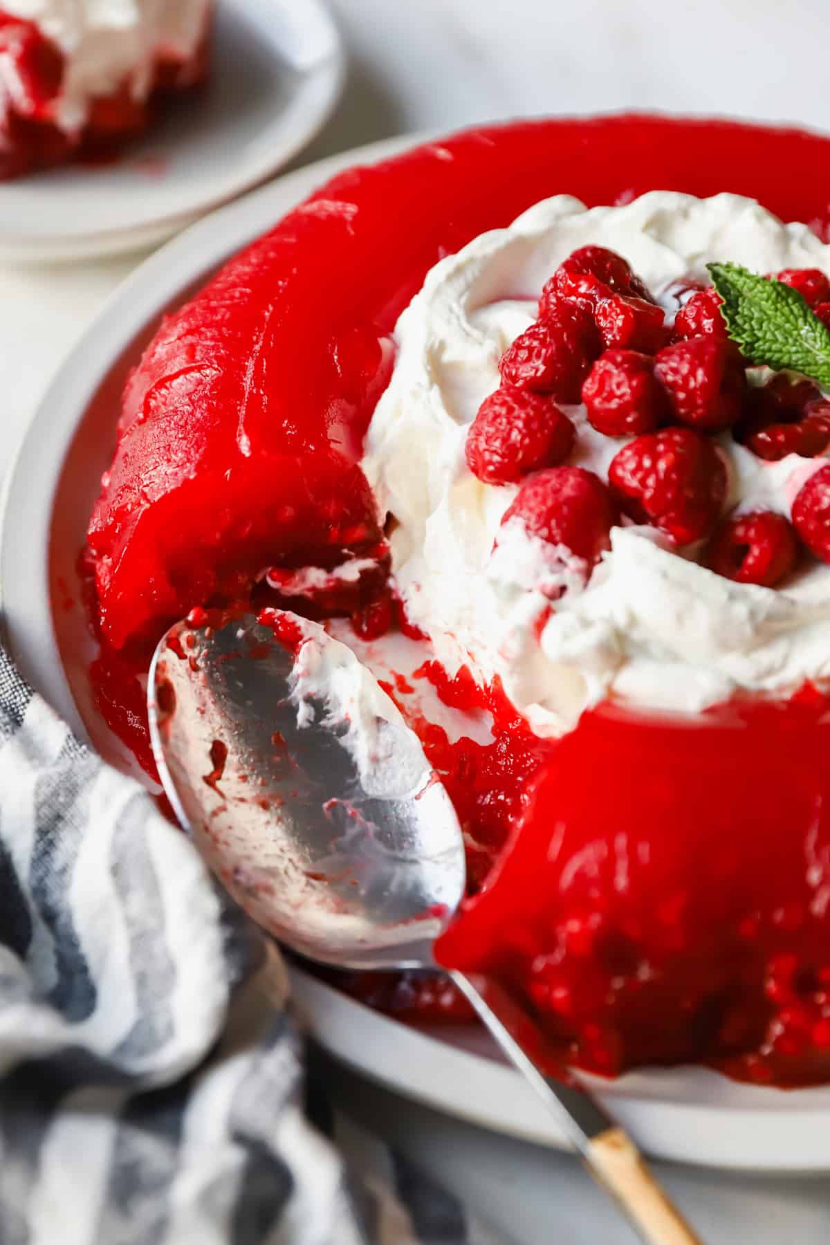 A red gelatin dessert topped with whipped cream and raspberries on a plate. A portion is missing, and a spoon is resting nearby.