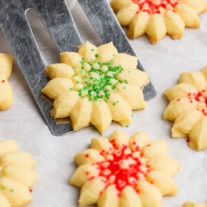 A spatula lifts a star-shaped, green-sprinkled cookie. Other cookies with red sprinkles are on a parchment-lined surface.