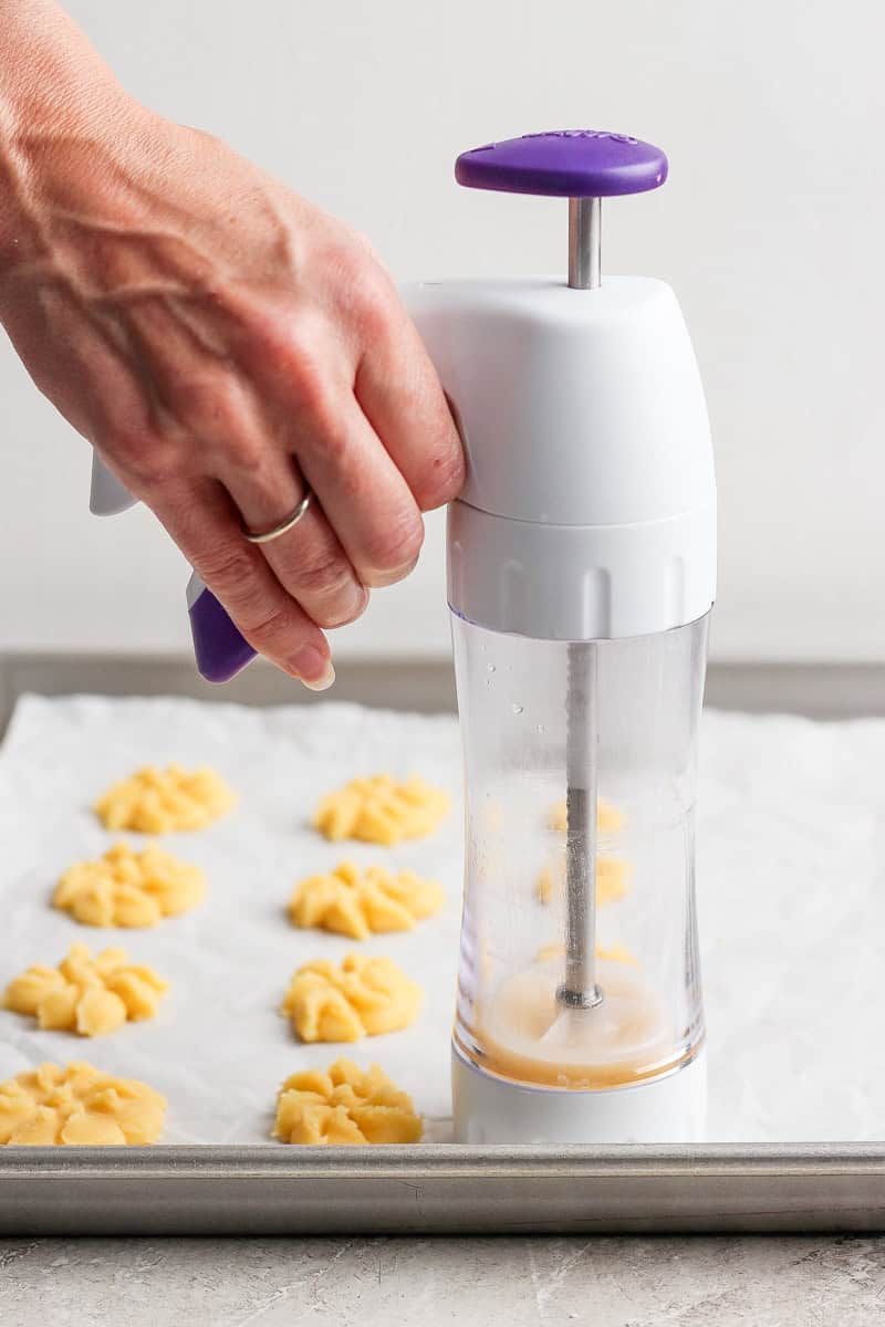 A hand pressing cookie dough onto a baking sheet using a cookie press, forming flower-shaped cookies.