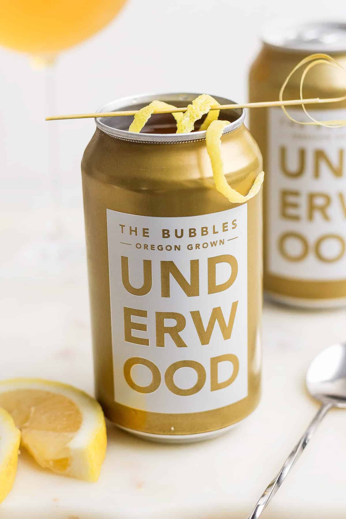 Underwood sparkling wine can with a lemon twist garnish sits next to lemon slices and a spoon on a white surface, creating an enticing canned champagne cocktail experience.