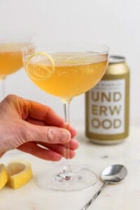 A hand holds a coupe glass filled with a fizzy canned champagne cocktail, garnished with a lemon slice. A can labeled "Underwood" and a spoon rest on a white surface, surrounded by lemon slices.