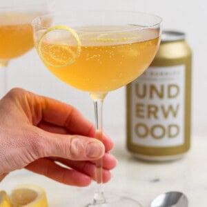 A hand holds a coupe glass filled with a fizzy canned champagne cocktail, garnished with a lemon slice. A can labeled "Underwood" and a spoon rest on a white surface, surrounded by lemon slices.