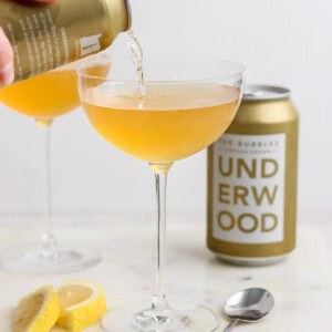 A can labeled "Underwood" is pouring a golden liquid, reminiscent of a canned champagne cocktail, into a coupe glass. Another filled glass, the can itself, lemon wedges, and a spoon are arranged on the marble surface.