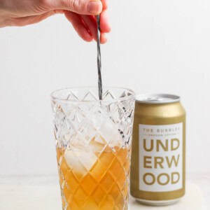 A hand stirs a canned champagne cocktail with ice in a diamond-patterned glass, while an unopened can of Underwood sparkling wine sits elegantly in the background.