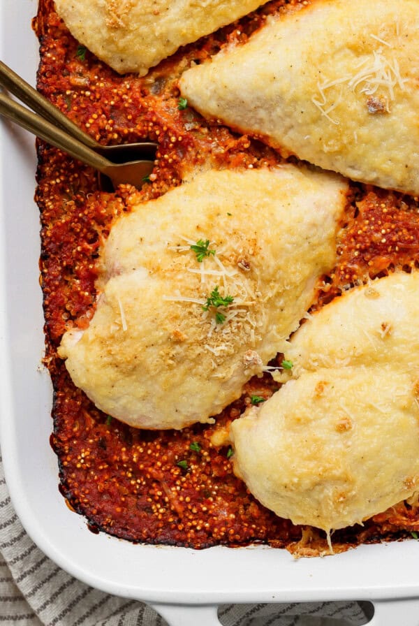 Baked chicken breasts topped with cheese on a bed of tomato quinoa in a white baking dish, garnished with herbs and cheese.