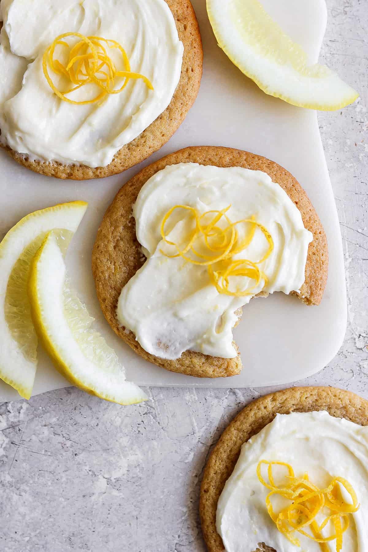 Three lemon cookies with cream cheese frosting, topped with fresh zest, sit invitingly on a cutting board. One is partially eaten, revealing its soft interior. Beside them, lemon wedges are artfully arranged on the textured surface, enhancing the citrusy allure of the scene.