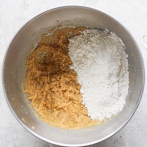 A mixing bowl with brown batter, white flour, and a hint of lemon zest sits on a gray surface, ready for transformation into delicious lemon cookies with cream cheese frosting.