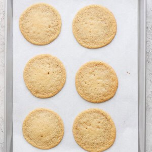 Six round lemon cookies with cream cheese frosting rest on a parchment-lined baking sheet.