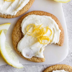 A lemon cookie with cream cheese frosting, topped with lemon zest, has a bite taken out of it. Slices of lemon are nearby on a light surface.