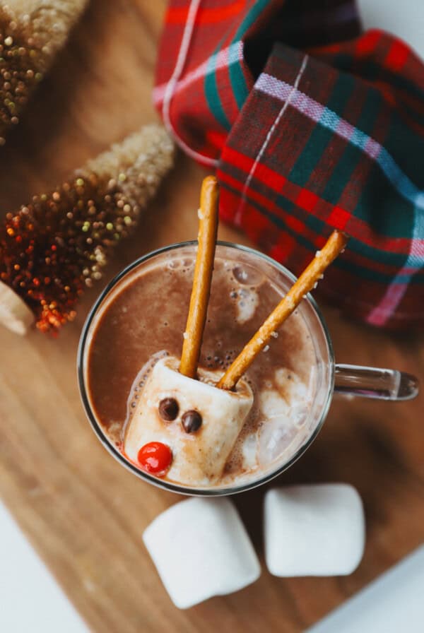 A cup of hot chocolate with a marshmallow decorated as a reindeer using pretzels, candy eyes, and a red candy nose, next to a plaid cloth and mini Christmas trees.