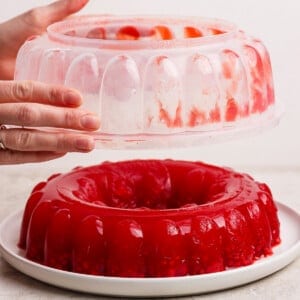 Hands removing a mold from a red gelatin dessert on a white plate.