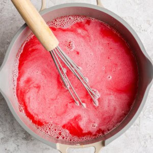 A whisk in a pot of frothy, red liquid on a light-colored surface.