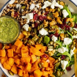 A bowl with spinach, wild rice, roasted butternut squash, walnuts, pepitas, cranberries, and feta cheese, served with a small container of pesto dressing.