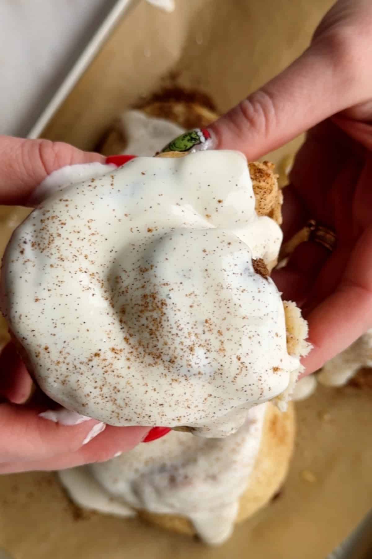 Close-up of hands holding a frosted protein cinnamon roll with a sprinkle of cinnamon on top.