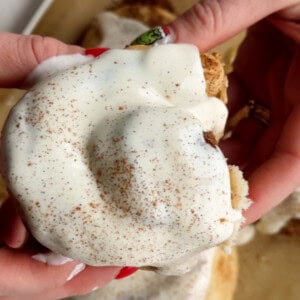 Hands holding a protein-infused cinnamon roll topped with cream cheese frosting and a sprinkle of cinnamon.